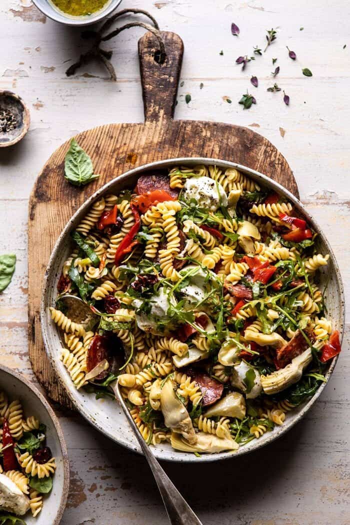 Antipasto Pasta Salad with Herby Parmesan Vinaigrette | halfbakedharvet.com #pasta #easyrecipes #pastasalad #mothersday Antipasto Pasta Salad with Herby Parmesan Vinaigrette with spoon in bowl and serving bowls next to pasta