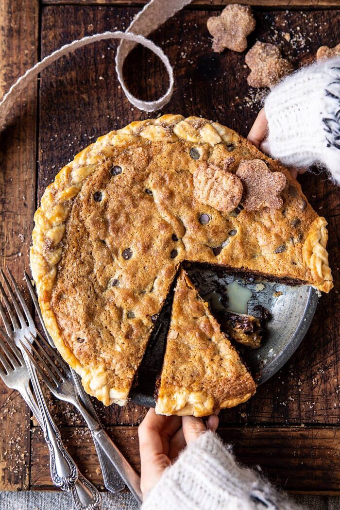 Gooey Chocolate Chip Cookie Pumpkin Pie | halfbakedharvest.com #cookiepie #thanksgiving #pumpkinpie overhead photo of Gooey Chocolate Chip Cookie Pumpkin Pie after baking with 2 hands on pie and 1 slice cut